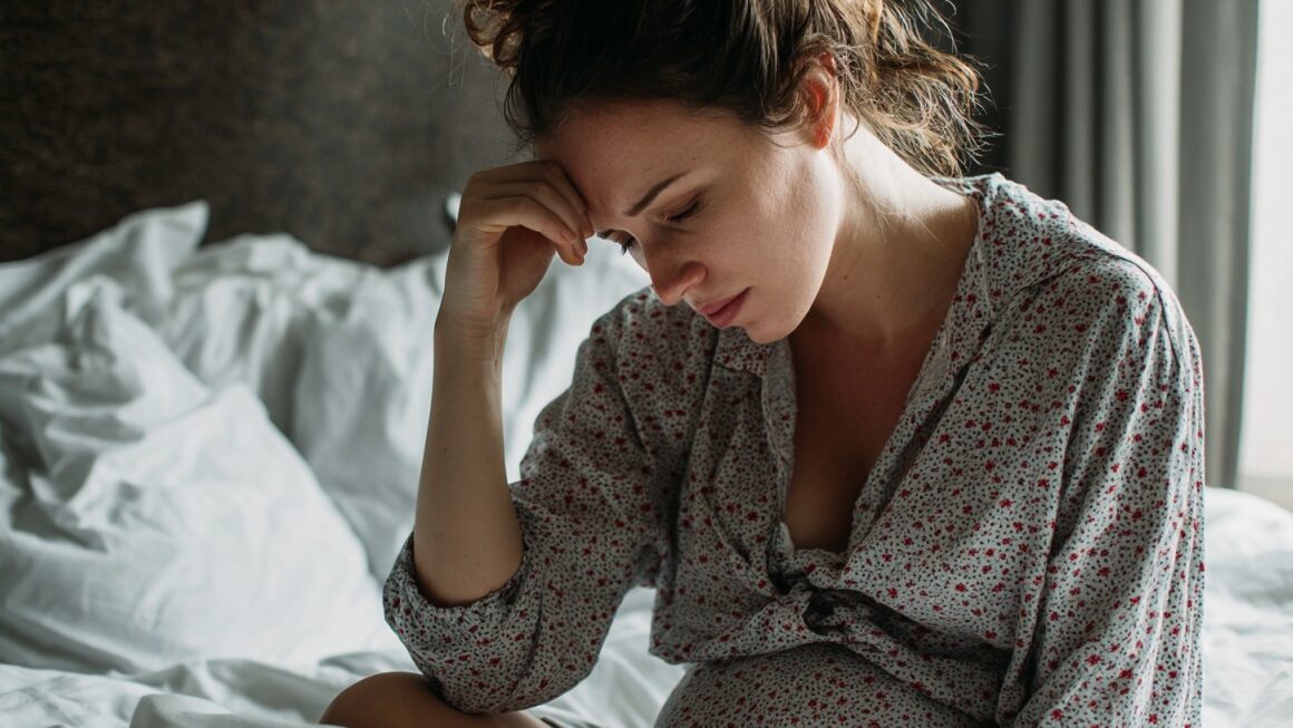 Trudnica sedi na krevetu i drži ruku na čelu zbog mučnine